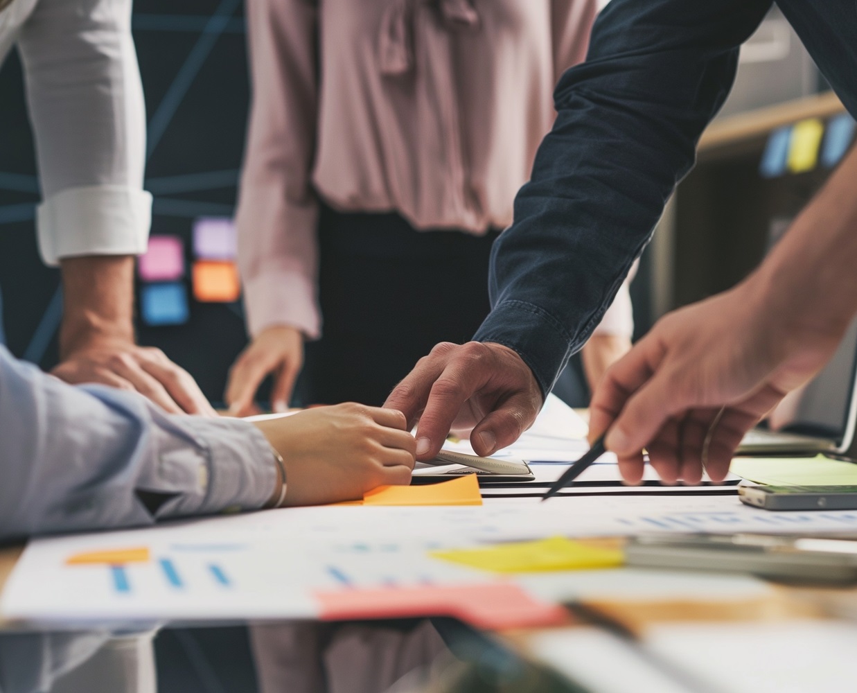 Close-up of a collaborative workspace with multiple people's hands pointing and working on documents with colorful sticky notes, charts, and a laptop on the table.