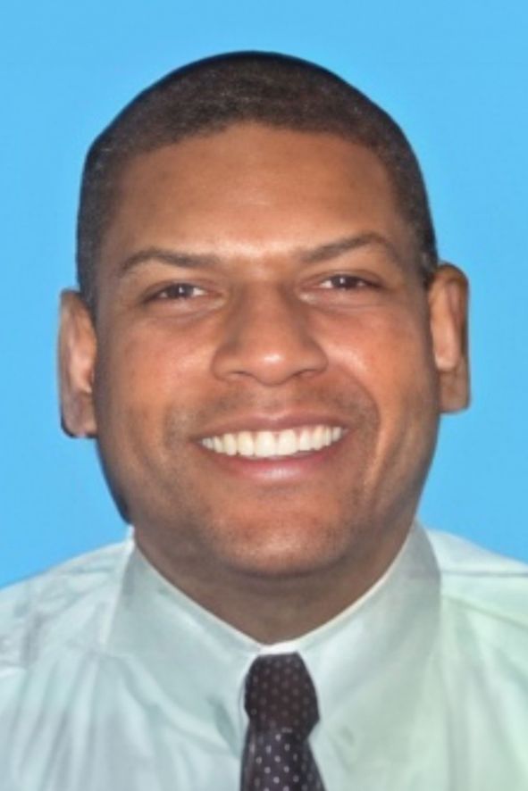 A portrait of a smiling man with short dark hair, wearing a light blue shirt and a dark polka dot tie against a solid blue background.
