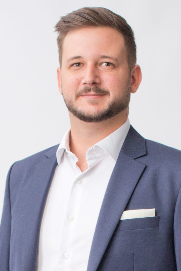 A man in a navy blue suit with a white shirt and pocket square, giving a slight smile against a plain background.