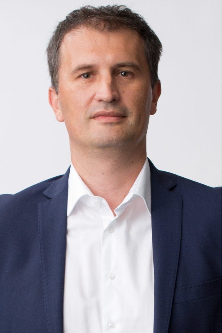 A portrait of a middle-aged man with short hair, wearing a dark blue suit jacket over a white button-up shirt, against a plain white background.