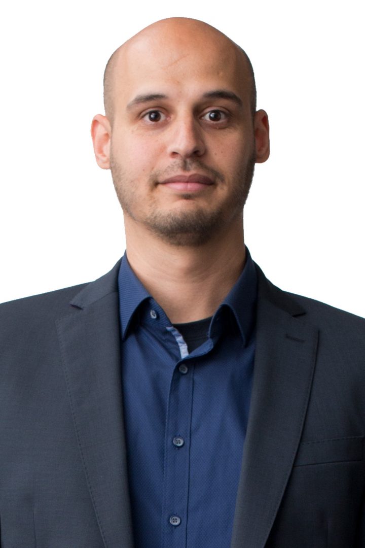 A bald man with a beard, wearing a dark gray suit jacket over a blue shirt, stands against a white background.