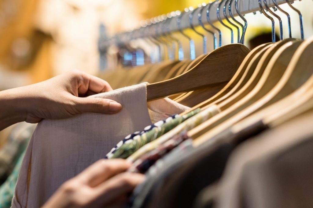 A person's hand browsing through a selection of clothes on wooden hangers in a store.