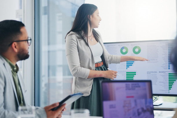 Woman in a light blazer pointing at a large monitor displaying pie charts and bar graphs during a presentation as a colleague with a tablet watches in a glass-walled conference room.