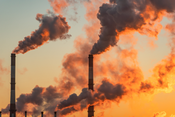 Multiple tall industrial chimneys releasing thick, dark smoke that billows and glows orange against a sunset sky.