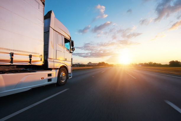 White semi-truck cab and trailer captured from the left side driving on a multi-lane highway toward a bright setting sun, with motion-blurred road and golden light reflecting off the truck under a partly cloudy sky.