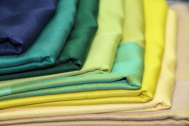 Close-up of neatly stacked folded fabrics arranged in a gradient from dark teal and greens to bright yellow and pale cream, showing smooth texture and a subtle twill weave along the folded edges.
